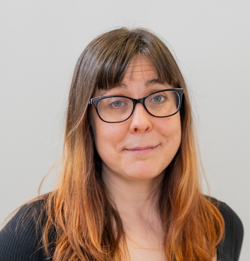 A photo of Emma. She is a white woman in her thirties with long brown hair that is slightly orange at the end. She has black glasses and blue eyes. She is wearing a black top