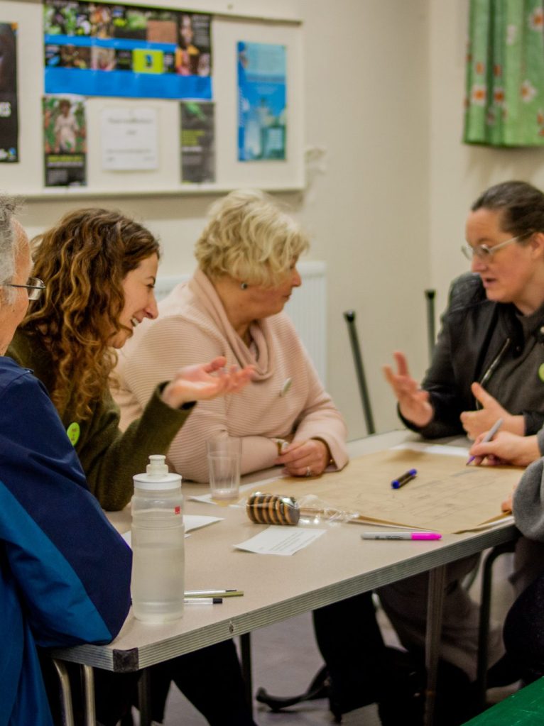 A group of women sit around a table, gesturing as they talk passionately to each other. Bottles and pens are on the table in front of them