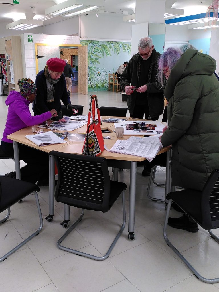 A group of older people sit or stand around a hexagonal table with papers laid out on it. They are discussing what parts to cut out