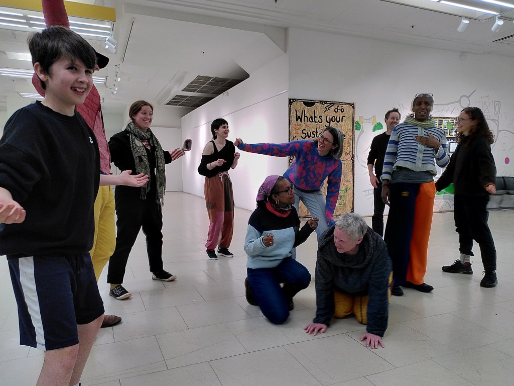 A group of people of all ages and backgrounds in dramatic poses in a large white room. They are smiling and interacting with each other