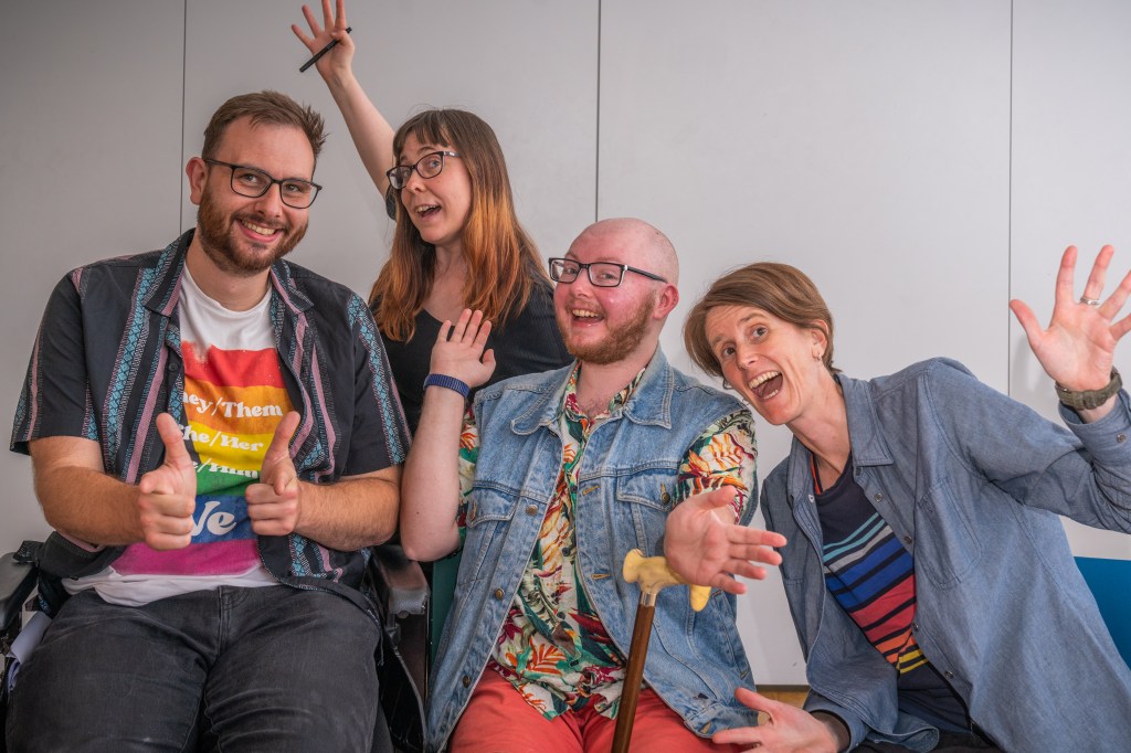 Four Disabled people wave at the camera while grinning and waving their arms