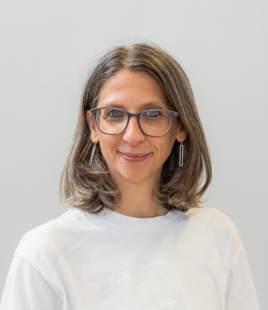 Rachel smiling at the camera. She is a white woman in her forties with shoulder length brown hair and glances. She is smiling at the camera and wearing dangling rectangular earrings
