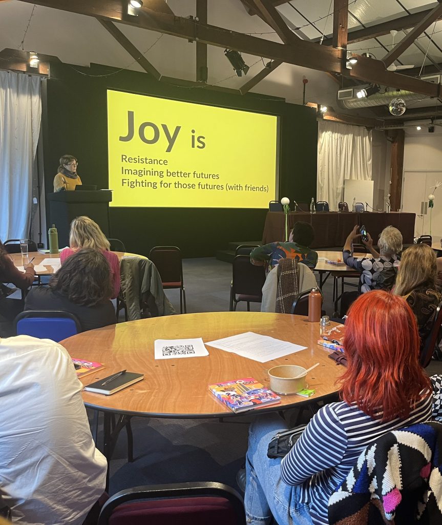 Emma speaking at the front of a room next to a slide show that is bright yellow and reads 'Joy is resistance, imagining better futures, fighting for those futures (with friends)' Lots of people sit around round tables listening