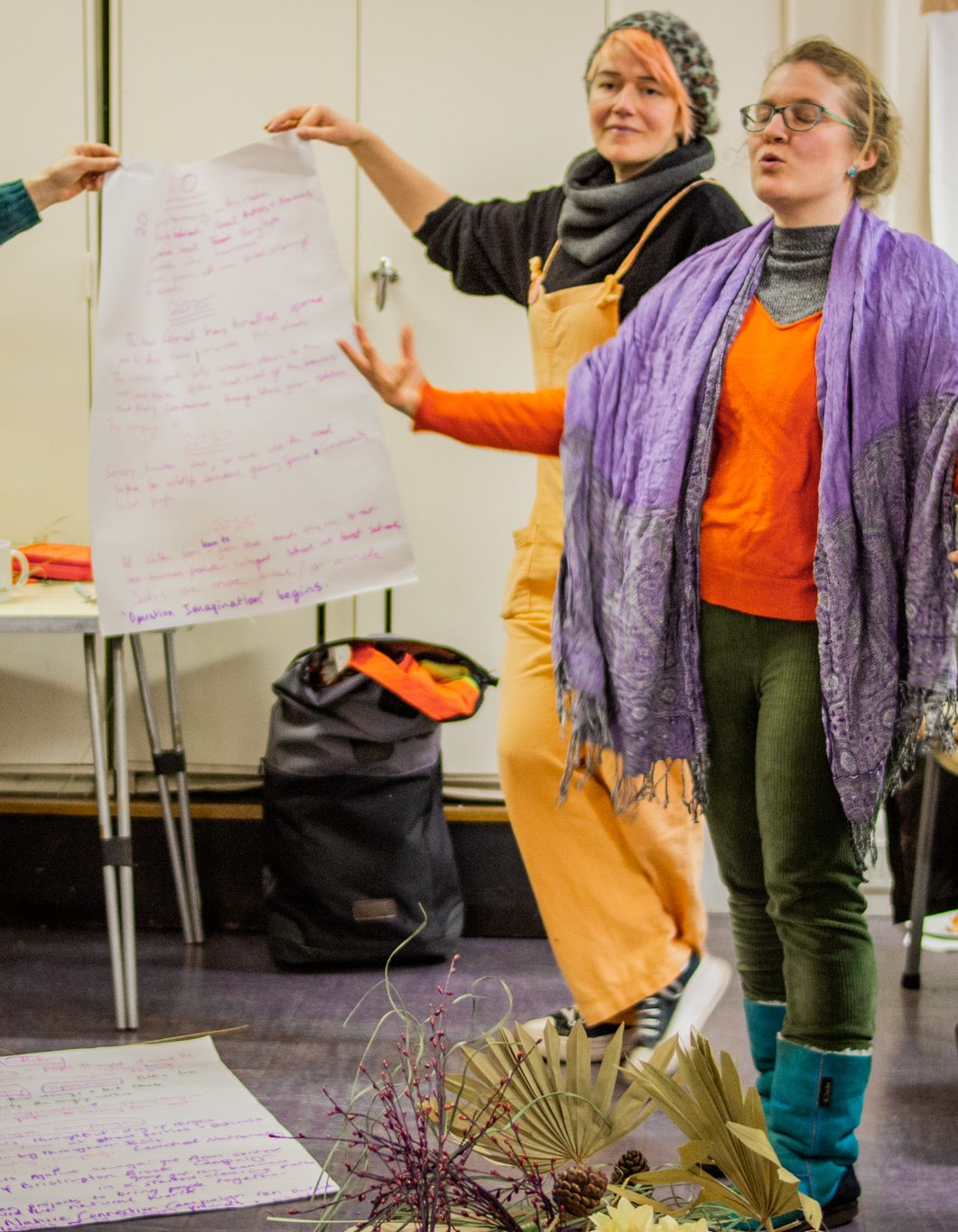 A group of women present a craft and large piece of paper with a story writeen on it. They are dressed in bright colours and the woman the front strikes a dramatic pose as she presents