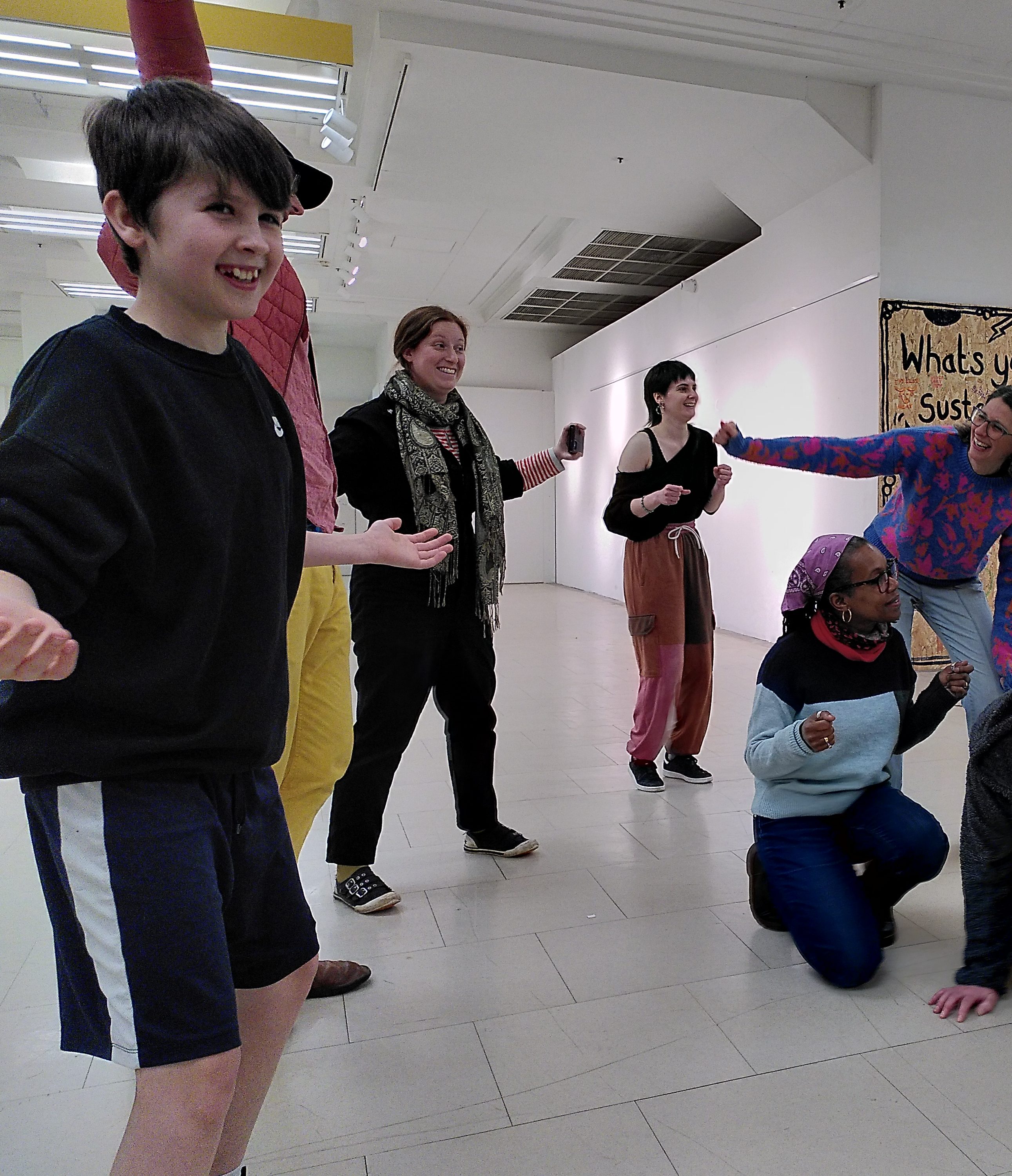 A group of people grin as they strike poses in a large space. Some people are standing, others are crouched or leant down. They are from all ages and backgrounds