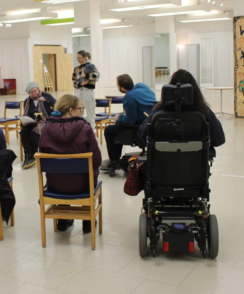 A group of Disabled people sit in a row watching two people preform a play. One person is sat down with a script, the other person is standing holding a fak baby